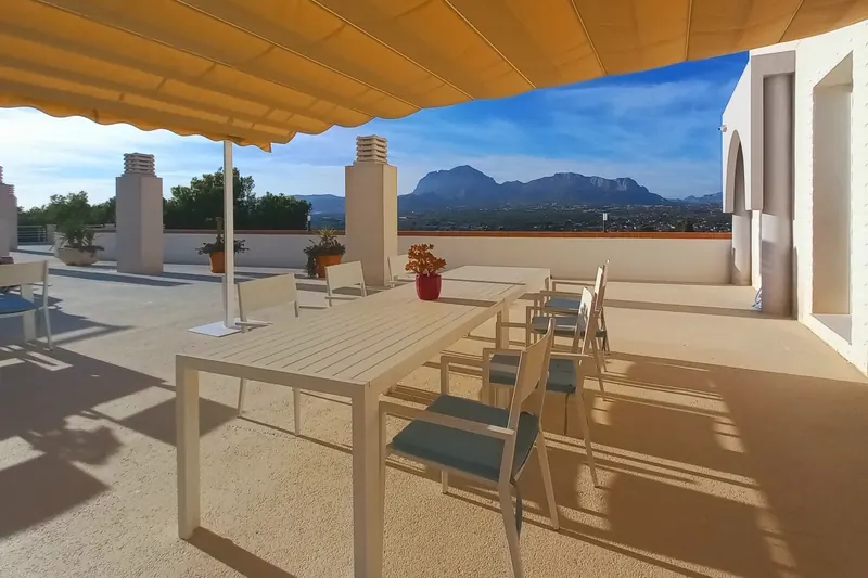 La azotea bajo un toldo amarillo: una mesa larga, sillas blancas con cojines gris azulado y la sierra al fondo.