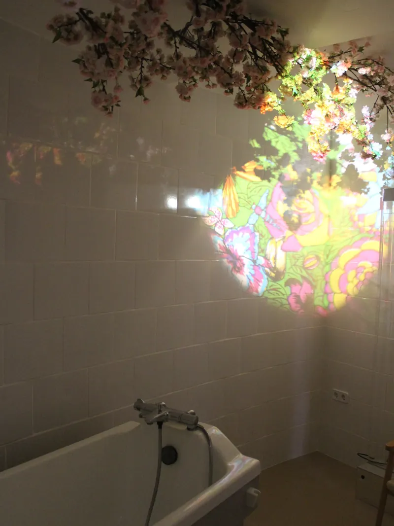 Un baño en penumbra con bañera, un dibujo de flores de colores proyectado en los azulejos y ramas con flores en el techo.