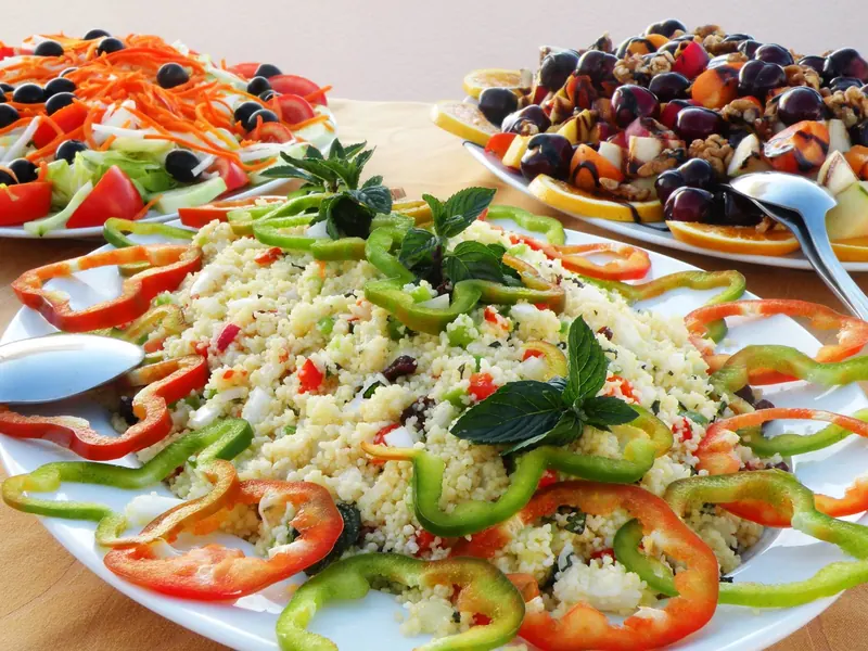 Una fuente de ensalada de cuscús con aros de pimiento rojo y verde y, detrás, una ensalada mixta y una fuente de fruta.