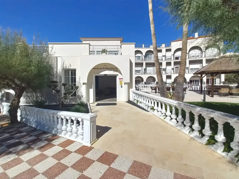 La entrada: un porche blanco en arco con el nombre de la residencia sobre la puerta, y un camino entre balaustradas blancas.