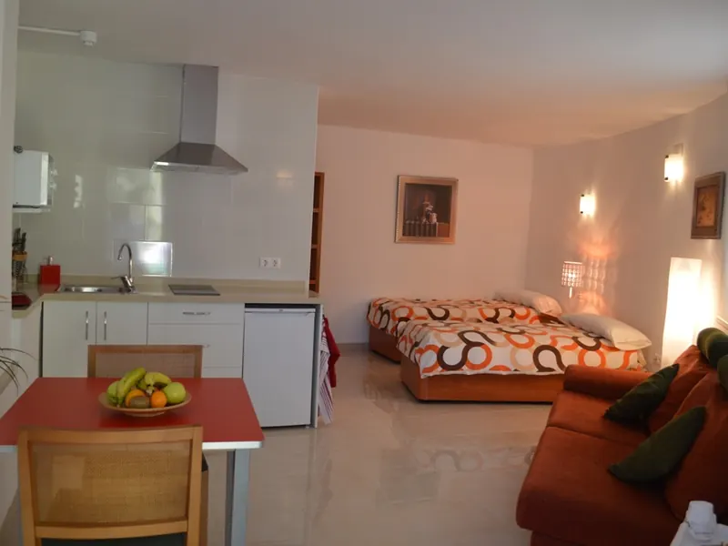 El estudio: una pequeña cocina, una mesa roja con un frutero, dos camas individuales y un sofá naranja.