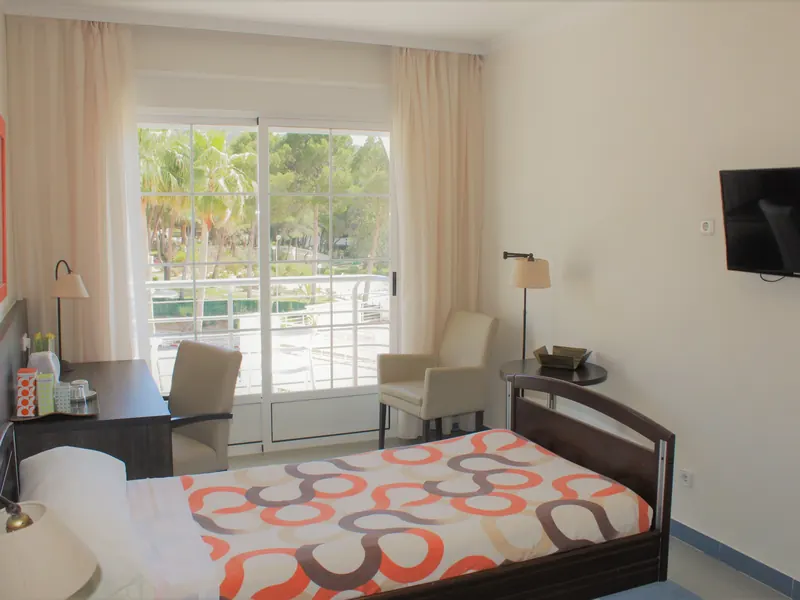 Una habitación: cama con colcha de dibujos naranjas, grises y marrones, sillón, televisor y una puerta acristalada al balcón, con palmeras detrás.