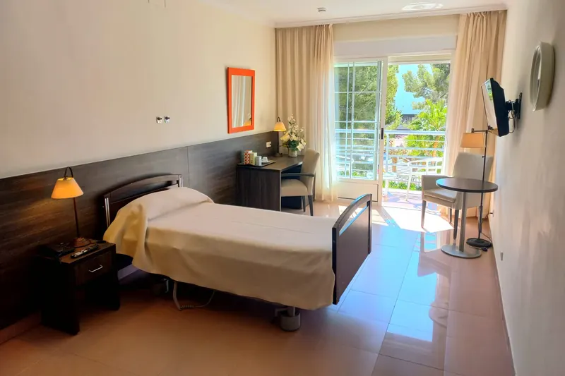Una habitación individual: cama regulable con cabecero de madera oscura, escritorio, televisor y una puerta acristalada abierta al balcón.