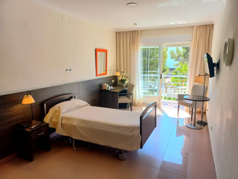 Una habitación individual: cama regulable con cabecero de madera oscura, escritorio, televisor y una puerta acristalada abierta al balcón.