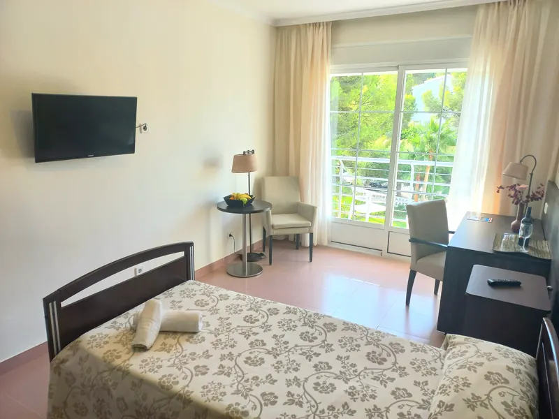 Una habitación: cama de madera oscura con colcha beis de flores, un televisor en la pared y una puerta de balcón con cortinas crema.