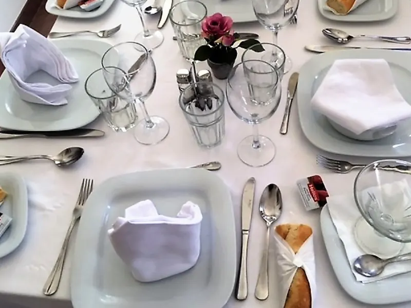 Una mesa puesta, vista desde arriba: platos cuadrados blancos con servilletas dobladas, copas, cubiertos, panecillos y una rosa.