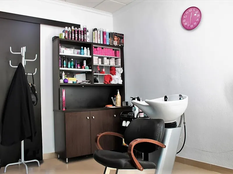 La peluquería: un sillón negro ante un lavacabezas blanco, un mueble con productos y toallas, y un reloj rosa en la pared.