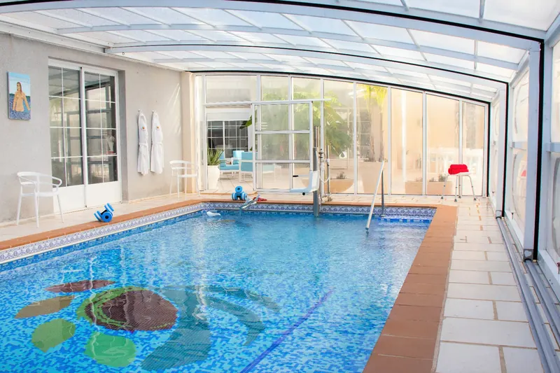 La piscina bajo una cubierta transparente: una tortuga de mosaico en el fondo, una silla elevadora y un pasamanos al otro lado.