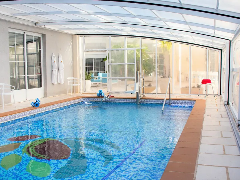 La piscina bajo una cubierta transparente: una tortuga de mosaico en el fondo, una silla elevadora y un pasamanos al otro lado.