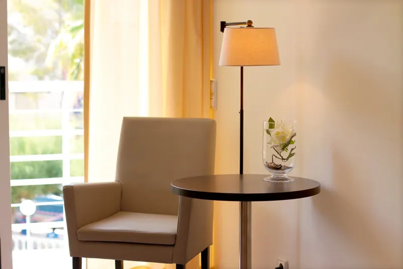Un rincón de una habitación: un sillón crema, una mesita redonda con una rosa blanca y una lámpara de pie encendida.