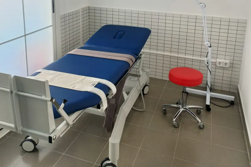 Una sala de tratamiento: una camilla azul regulable sobre ruedas y un taburete rojo.