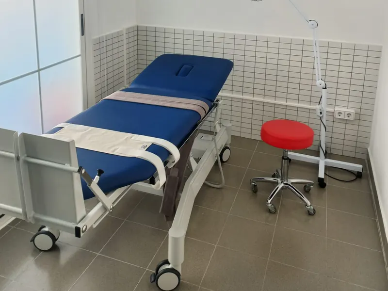 Una sala de tratamiento: una camilla azul regulable sobre ruedas y un taburete rojo.