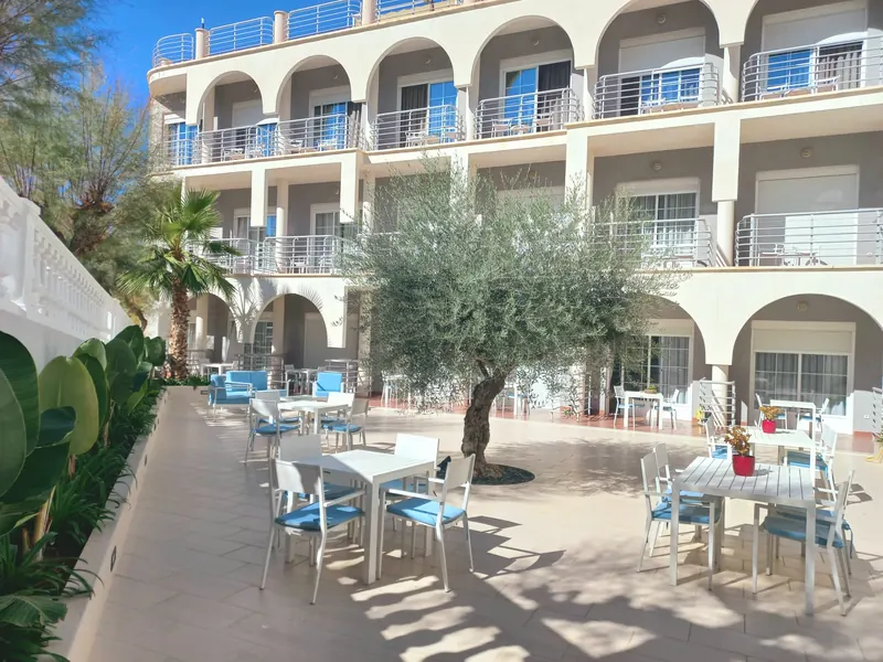 La terraza ante el edificio de balcones en arco: un olivo en el centro y mesas blancas con sillas de cojines azules.